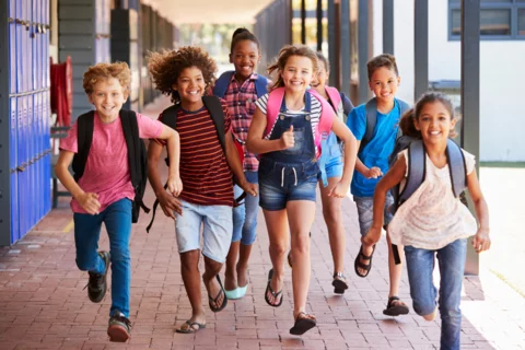 Photograph of a group of 7 smiling kids running down the hallway of their school with backpacks towards the camera.