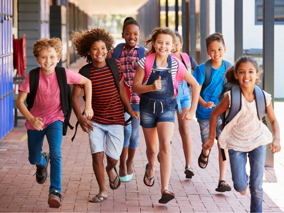 Photograph of a group of 7 smiling kids running down the hallway of their school with backpacks towards the camera.