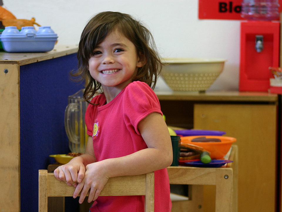 Photograph of a girl smiling at the camera while looking at the camera in a school classroom.